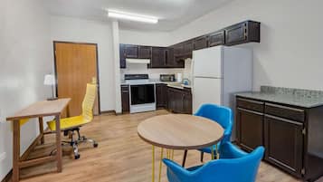Una cocina compacta con armarios de madera oscura, un refrigerador blanco, un microondas y una estufa. Hay una pequeña mesa de comedor con dos sillas, una amarilla y otra azul.