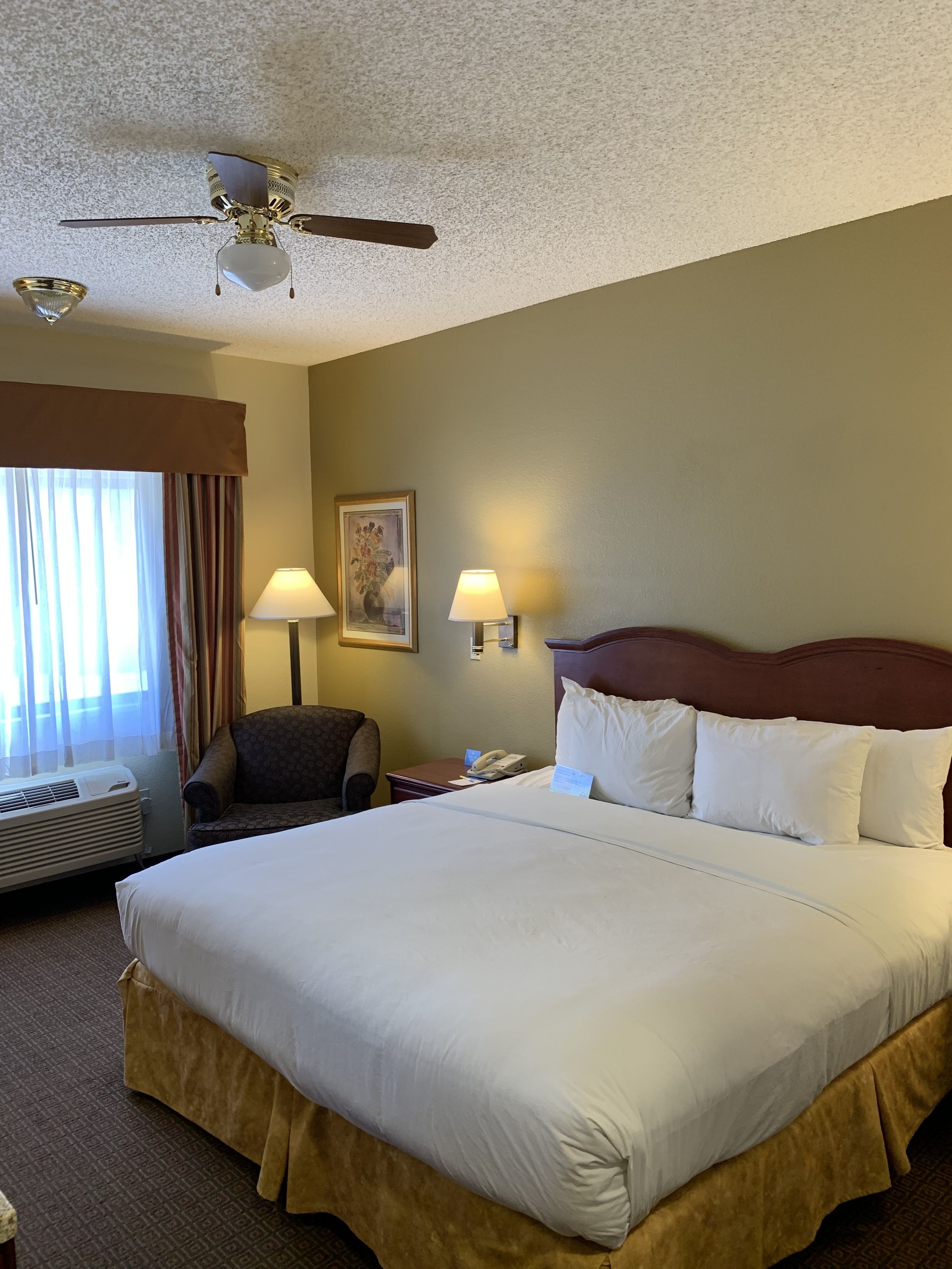 A hotel room with a large bed, two bedside lamps, a ceiling fan, a chair, a desk with a phone, and a window with curtains.