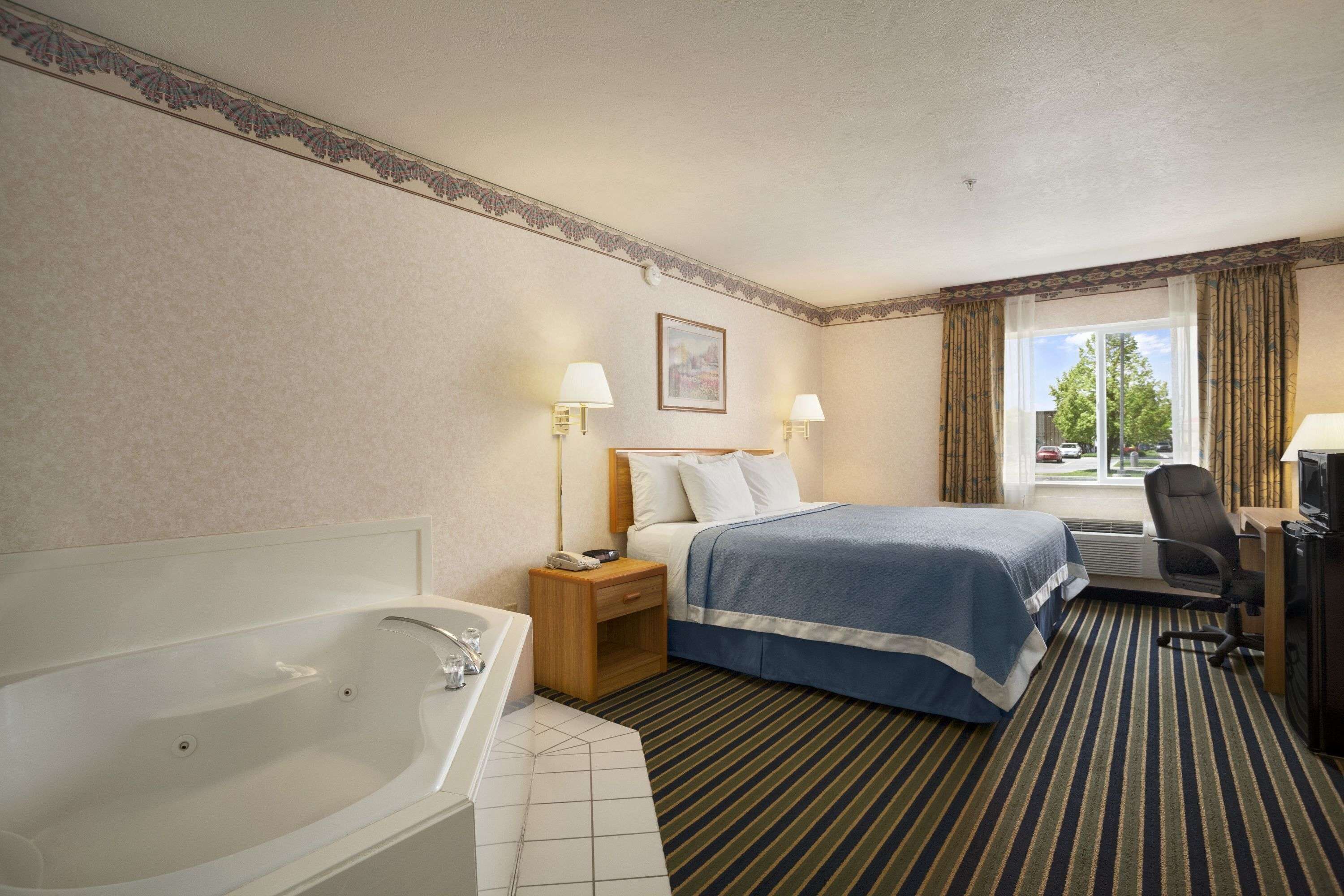 Ein Hotelzimmer mit einer großen Badewanne, einem Bett mit blauer Bettdecke, zwei Nachttischlampen, einem Schreibtisch mit Computer und einem Fenster mit Vorhängen.