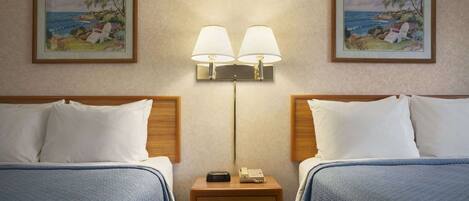 A hotel room with two beds, a nightstand with a lamp, and two framed coastal paintings on the walls.