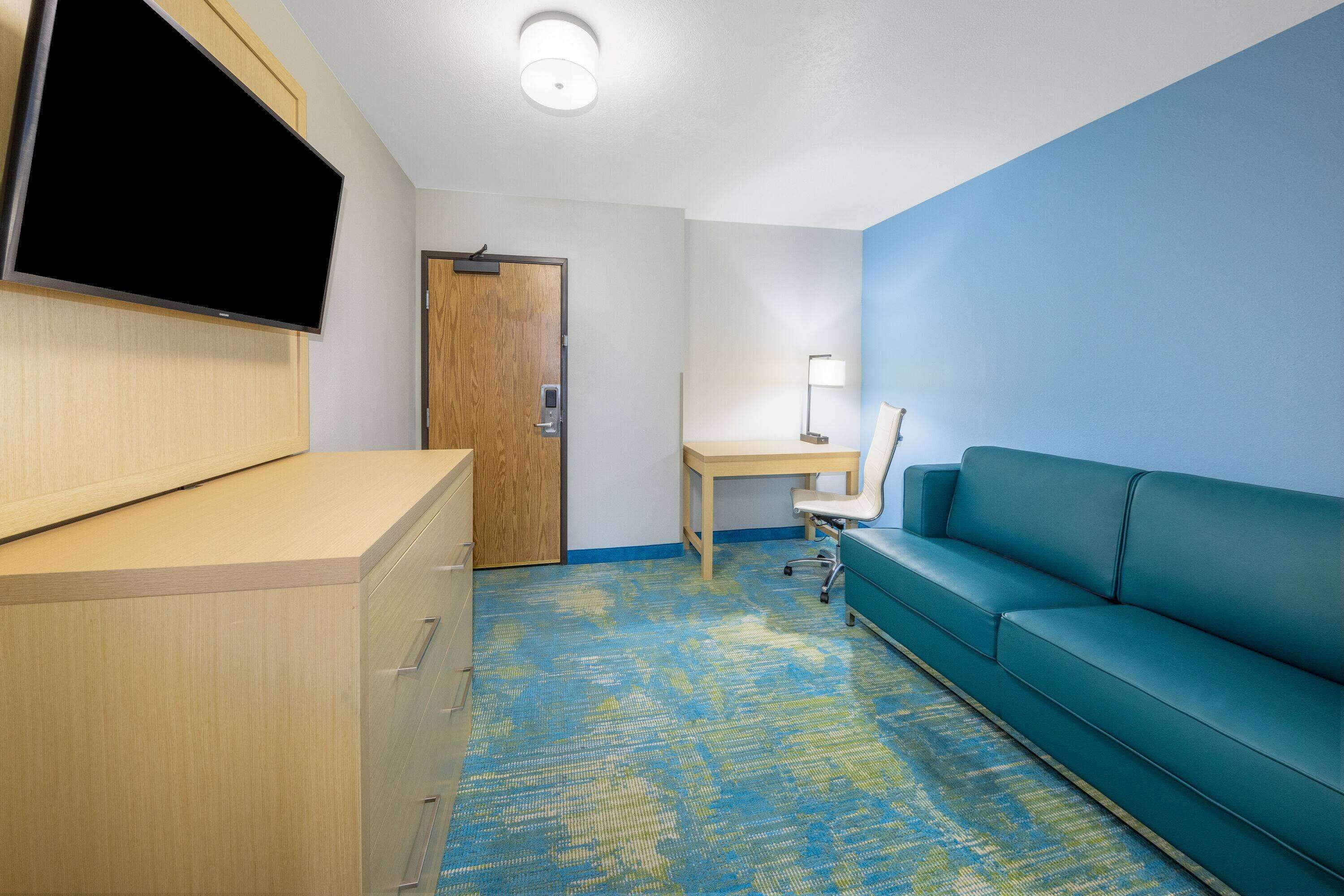 Habitación de hotel con escritorio, sofá azul, televisor montado en un mueble y alfombra estampada.
