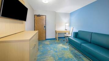 Habitación de hotel con escritorio, sofá azul, televisor montado en un mueble y alfombra estampada.