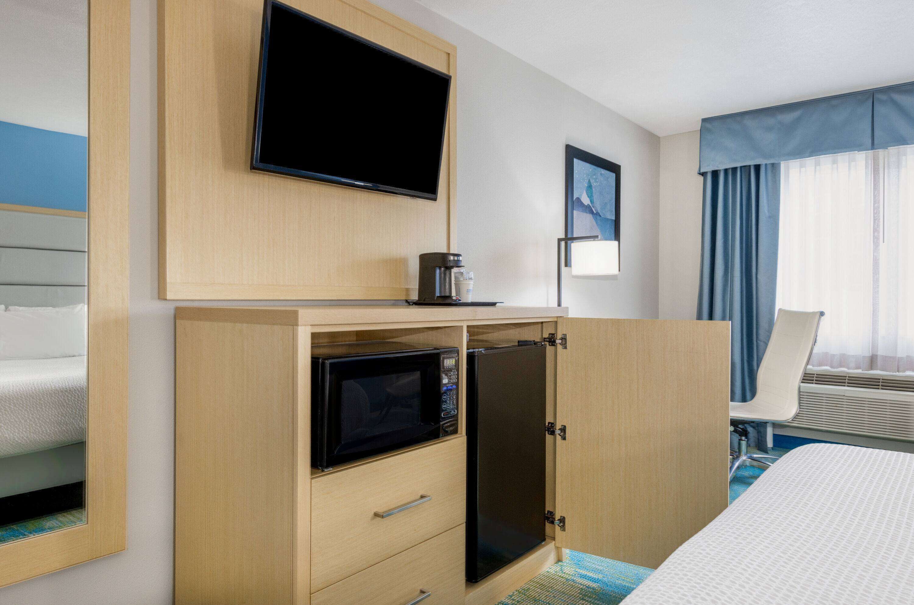 A hotel room with a bed, a flat-screen TV mounted on a wooden cabinet, a microwave, a small refrigerator, a bedside table with a lamp, and a window with curtains.