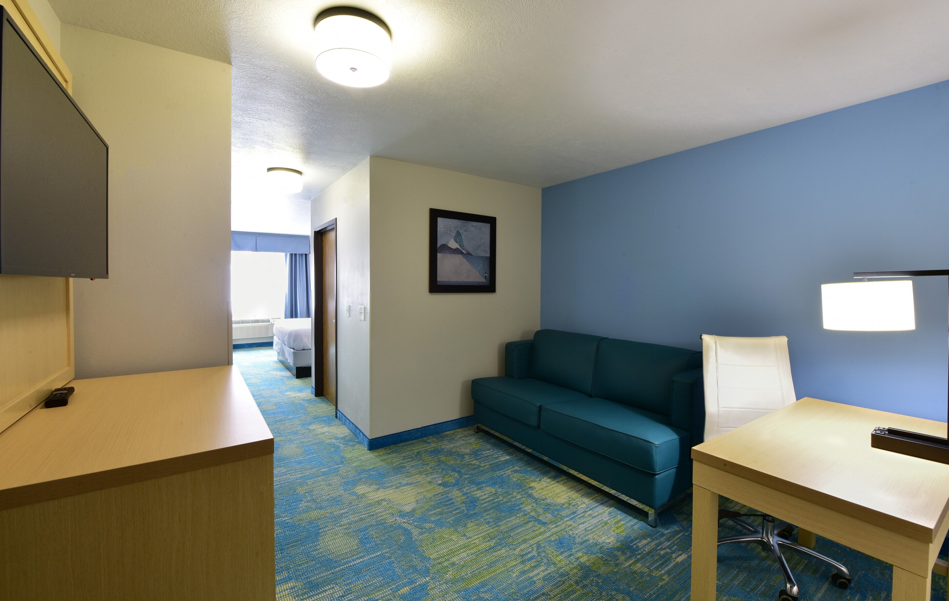 A hotel room with a desk, a couch, a TV, and a bedside lamp.