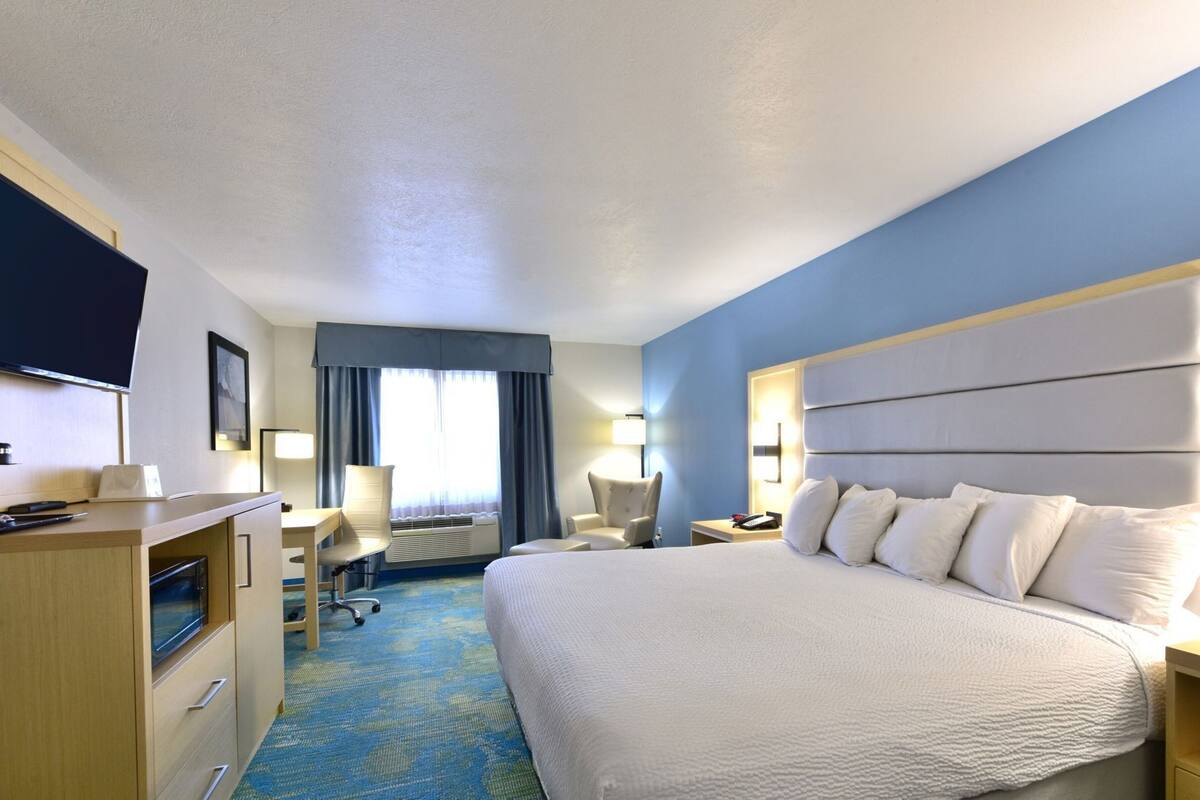 A hotel room with a large bed, a desk with a computer, a TV, and a window with curtains.