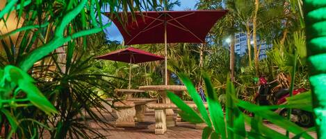 An outdoor seating area with stone tables and red umbrellas, surrounded by palm trees and a brick pathway.