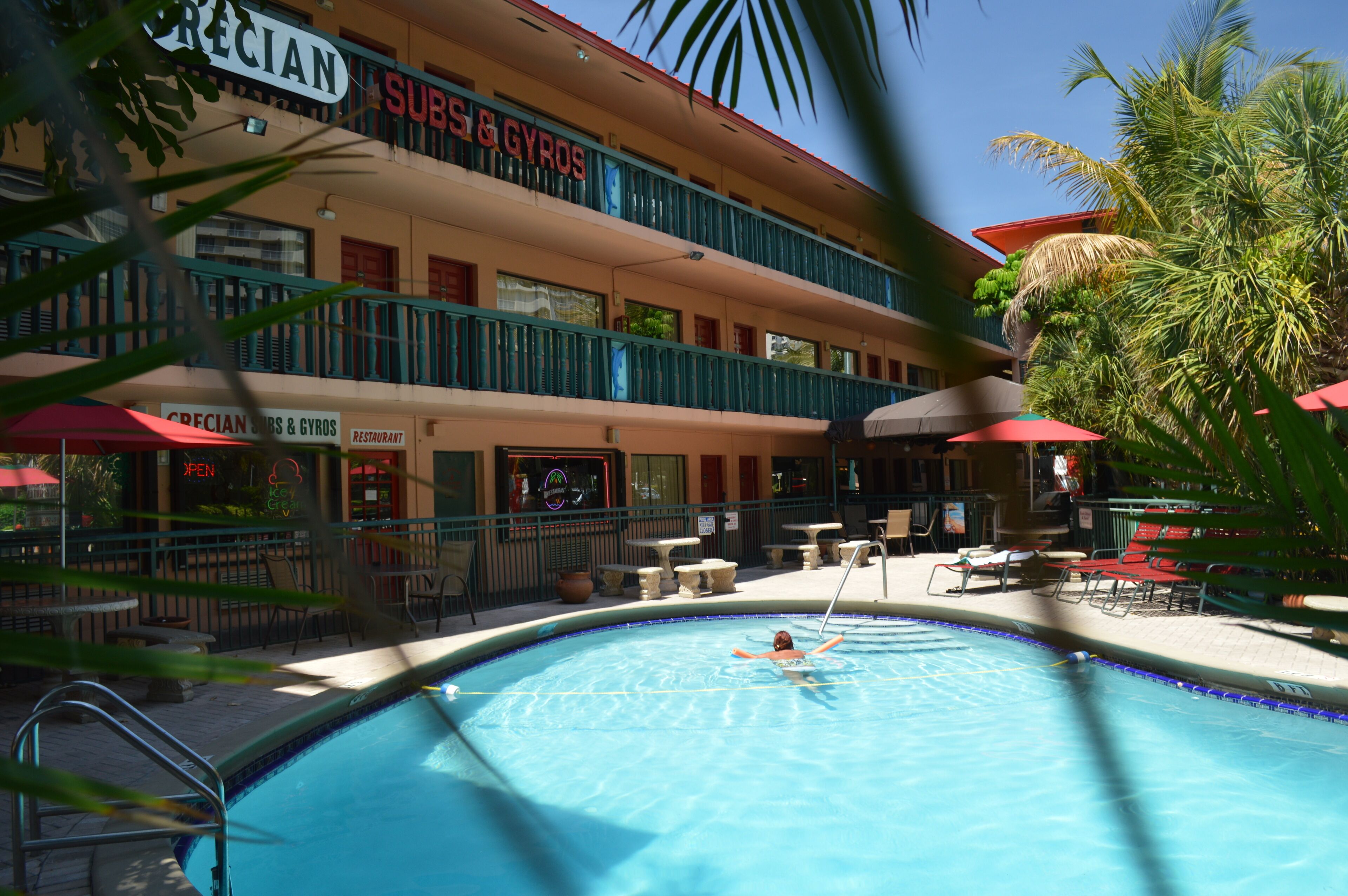 Une piscine où une personne nage, entourée d’espaces de détente en plein air et d’un restaurant appelé « CRECIAN SUBS & GYROS ».