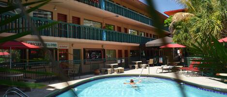 A swimming pool with a person swimming, surrounded by outdoor seating and a restaurant named "CRECIAN SUBS & GYROS".