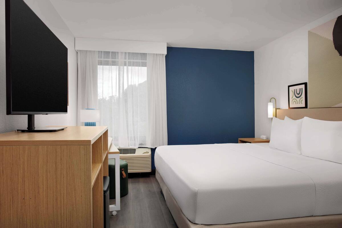 Et hotelværelse med en stor seng, et skrivebord med et fladskærms-tv, et sengebord med en lampe og et vindue med lette gardiner.