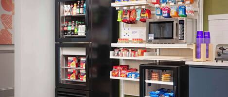 A kitchen area with a refrigerator stocked with beverages and snacks, a microwave, and shelves with packaged food items.