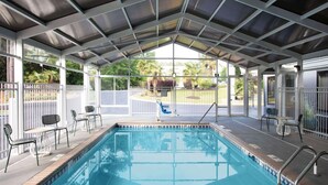 Una piscina interior con techo cubierto, rodeada de sillas y mesas, y vistas al jardín exterior.