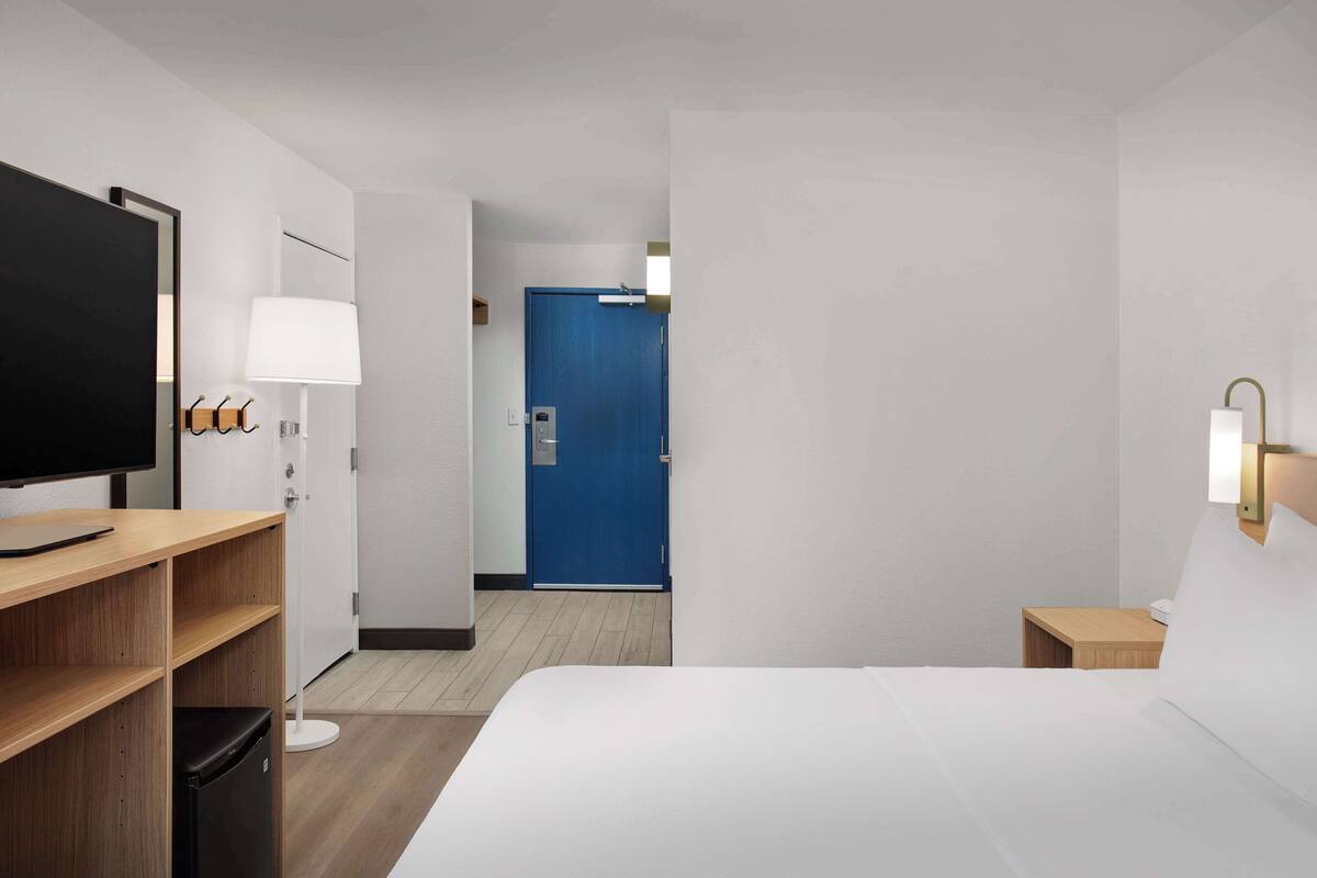 Et hotelværelse med en seng, et fjernsyn, et skrivebord med lampe, et lille natbord og en blå dør i baggrunden.