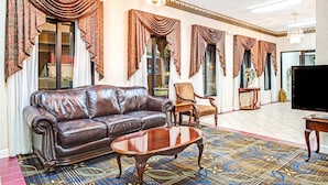 Amplia sala de estar con sofá de cuero, mesa de centro de madera y televisión. La habitación tiene ventanas grandes con cortinas ornamentadas y alfombra estampada.