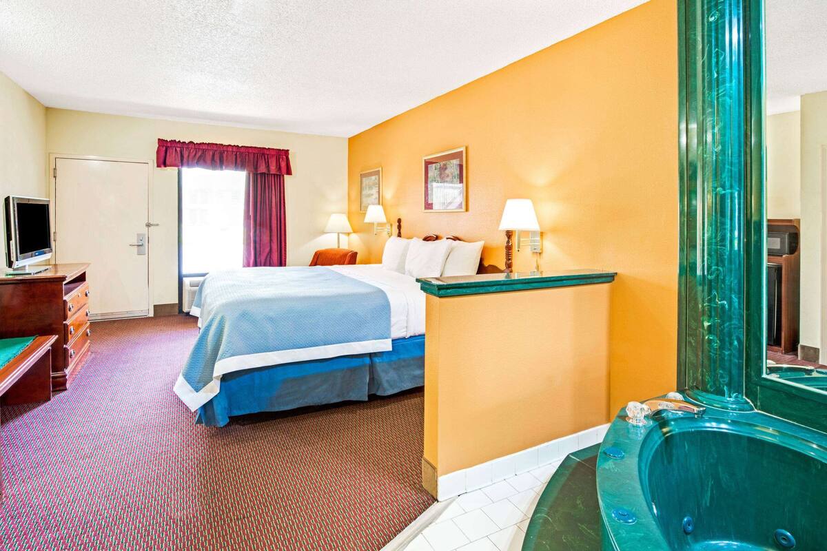 A hotel room with a bed, a desk with a computer, a TV, a bathroom with a bathtub, and a window with curtains.
