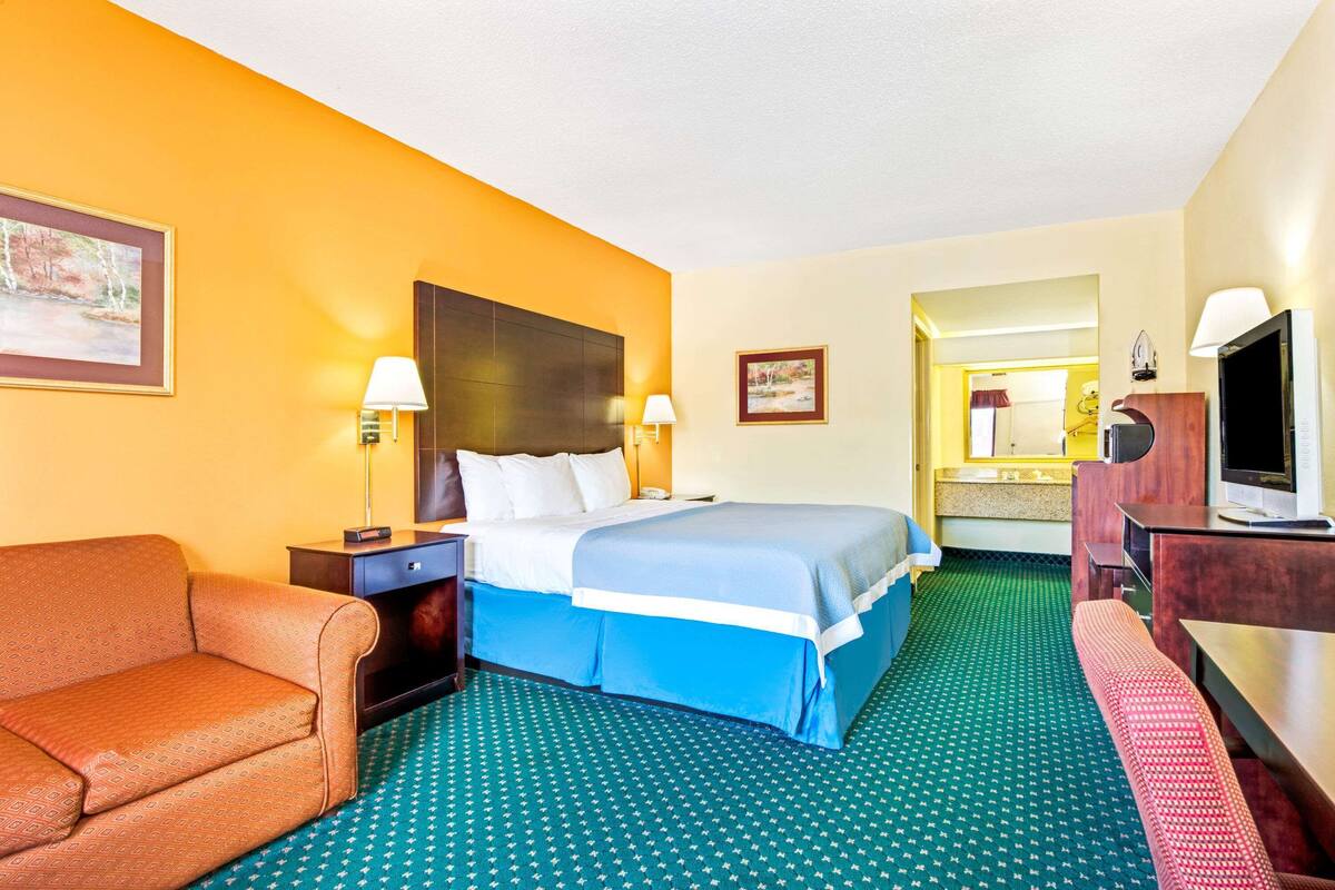 A hotel room with a bed, a sofa, a desk, a TV, and a bathroom visible through an open door.