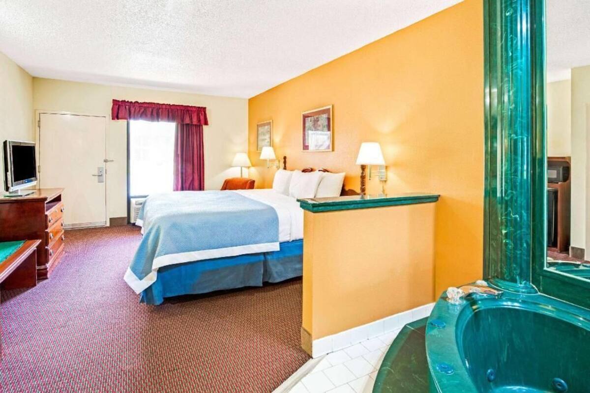 A hotel room with a bed, bedside lamps, a TV, a desk, a dresser, a window with curtains, and a jacuzzi.