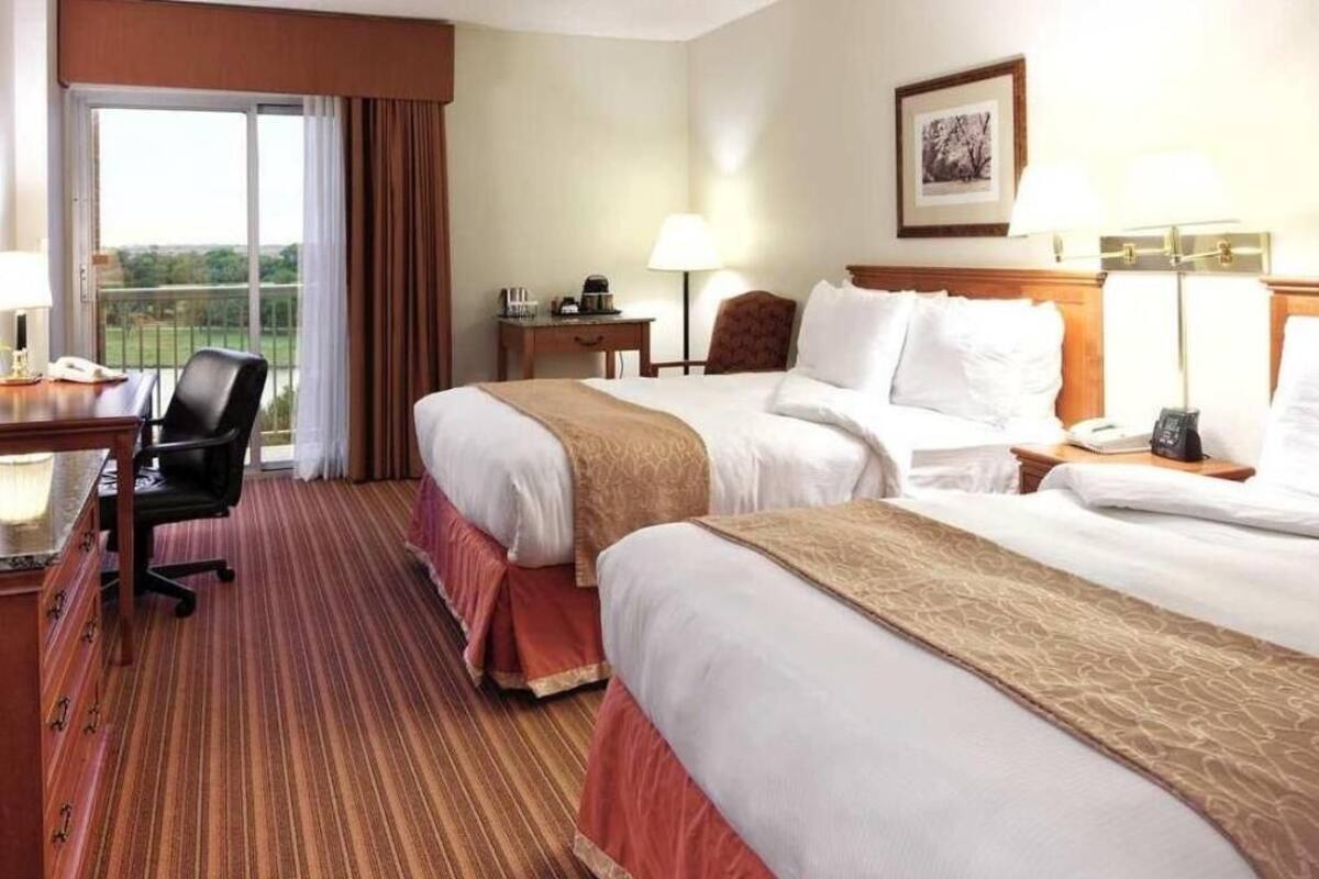 A hotel room with two beds, a desk, a chair, a lamp, and a window with curtains.