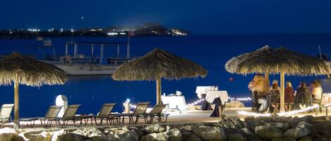 A beachside dining area with thatched umbrellas, lounge chairs, and a view of the sea and distant island.