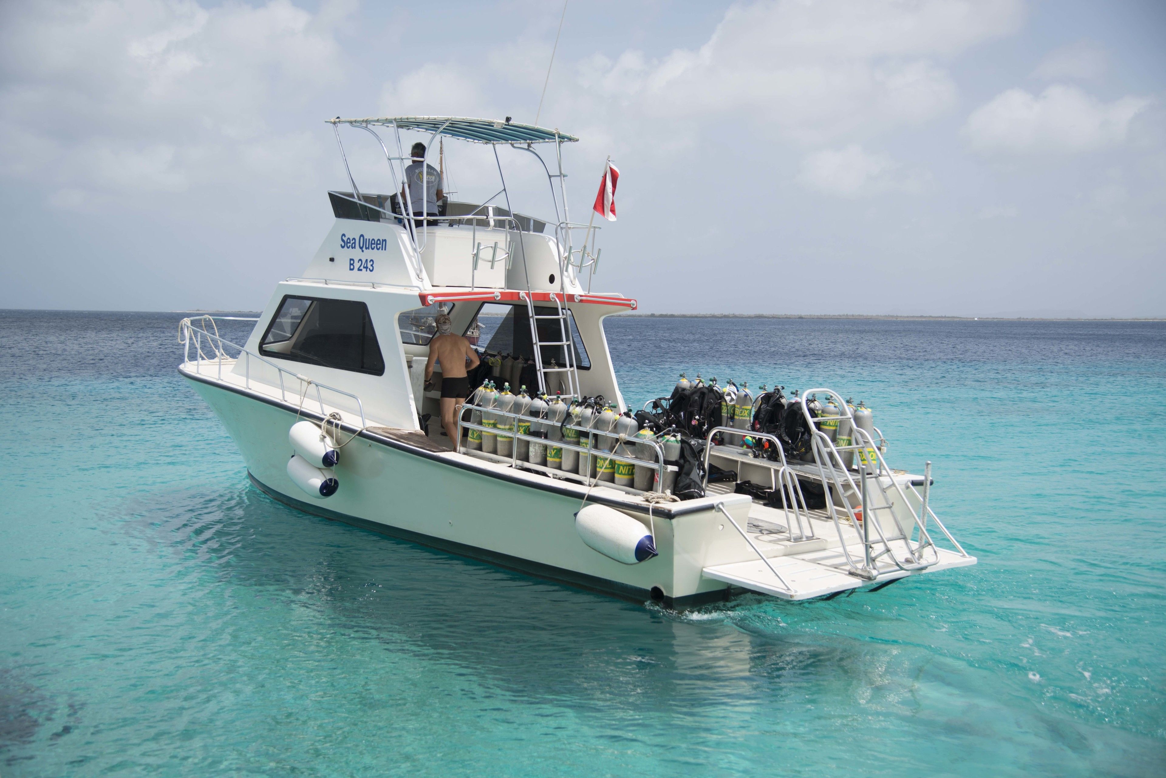 Una embarcació blanca anomenada 'Sea Queen' amb una bandera, equipada amb material de submarinisme i una escala, flotant sobre aigües turqueses cristal·lines.