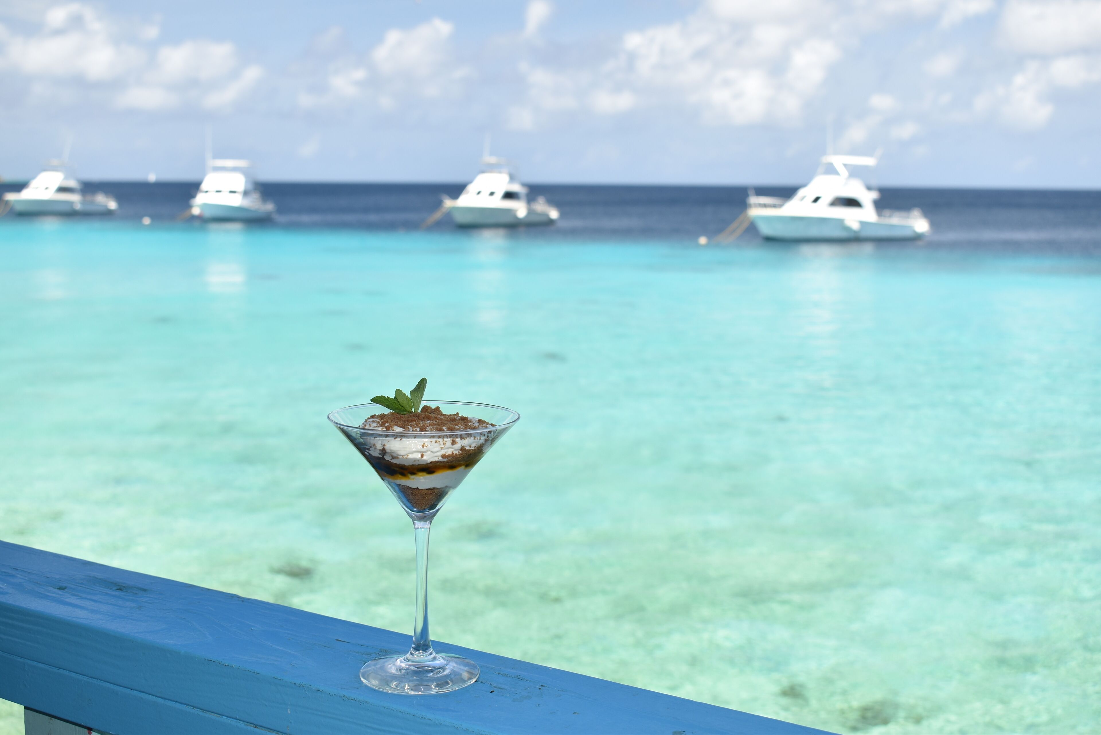 Una copa de martini con un postre sobre una barandilla azul, con vista al mar turquesa y barcos.
