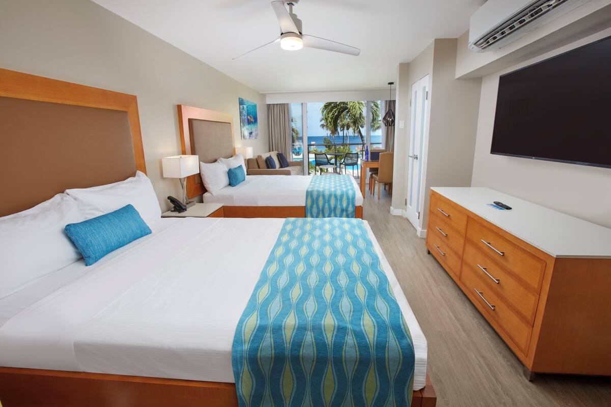 Habitación de hotel con dos camas, televisión, escritorio y balcón con vista al océano.