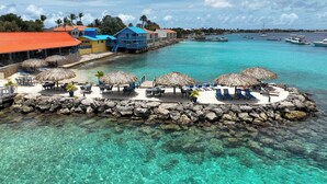 Een tropisch strandresort met helder turquoise water, rieten parasols en diverse accommodaties, waaronder een blauw gebouw met balkon.