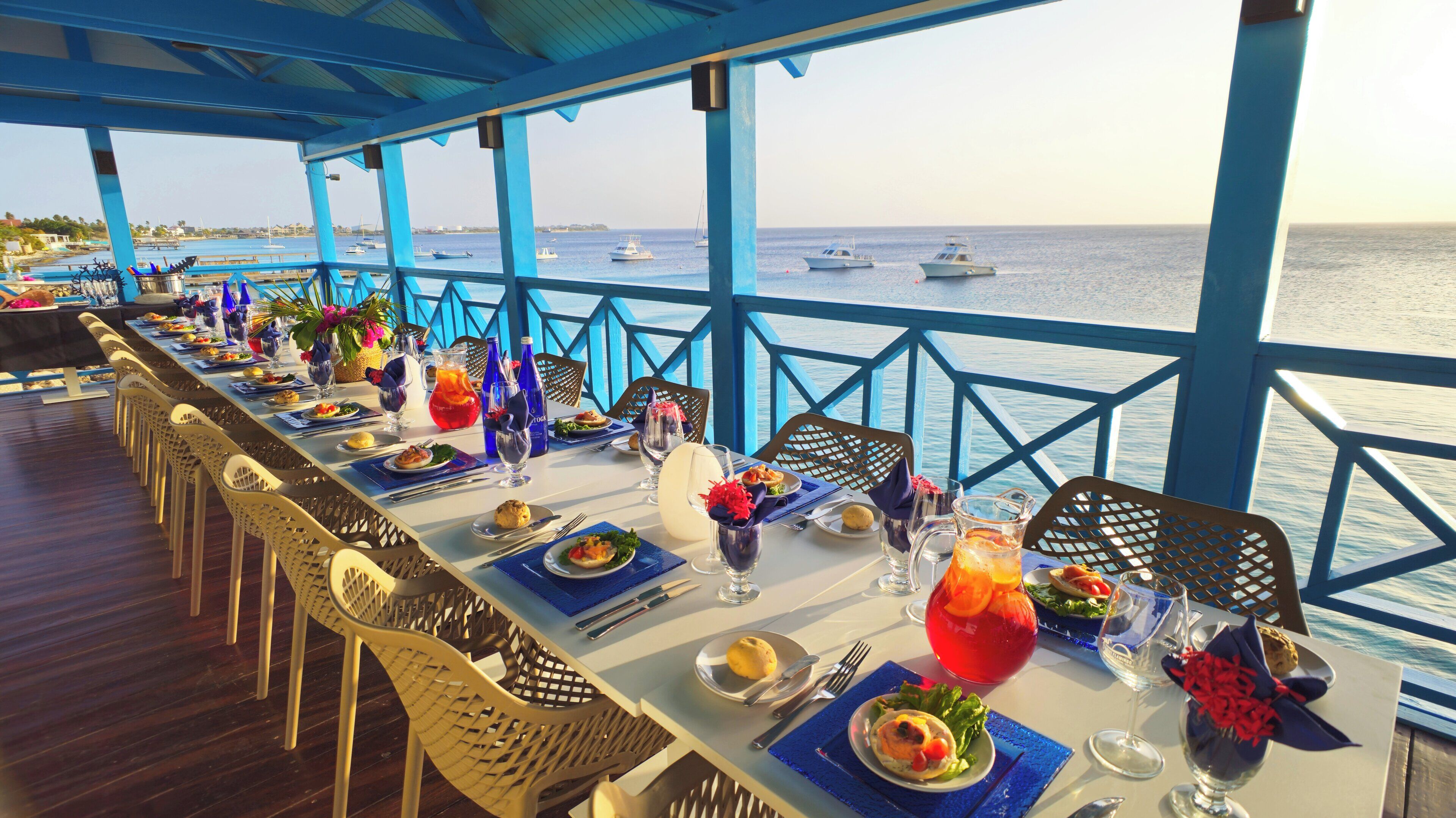 A long dining table set with plates, cutlery, and drinks, overlooking a body of water with boats.