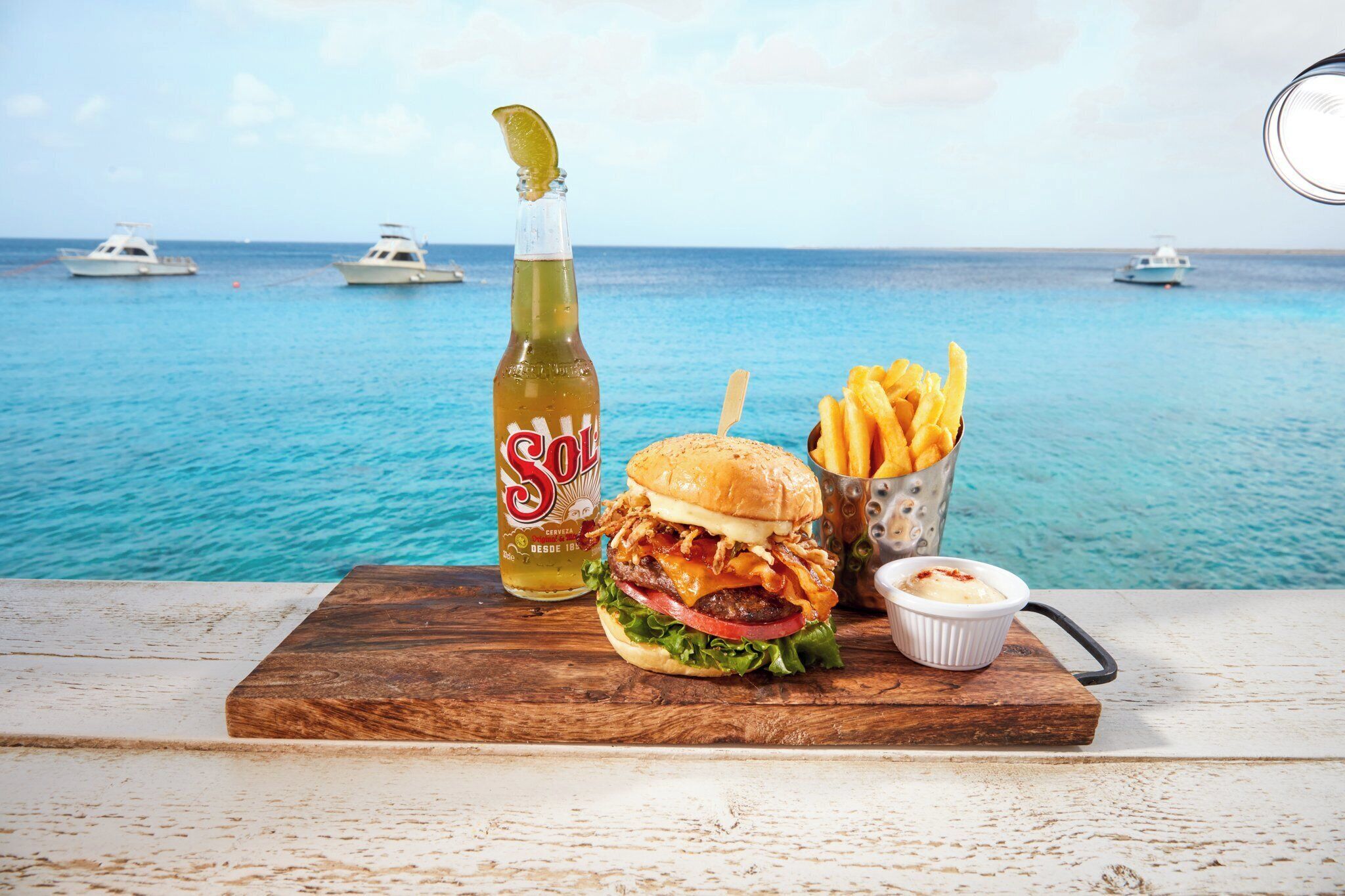 Ein Essen mit einem Burger, Pommes und einem Getränk vor einer Strandkulisse mit Booten.