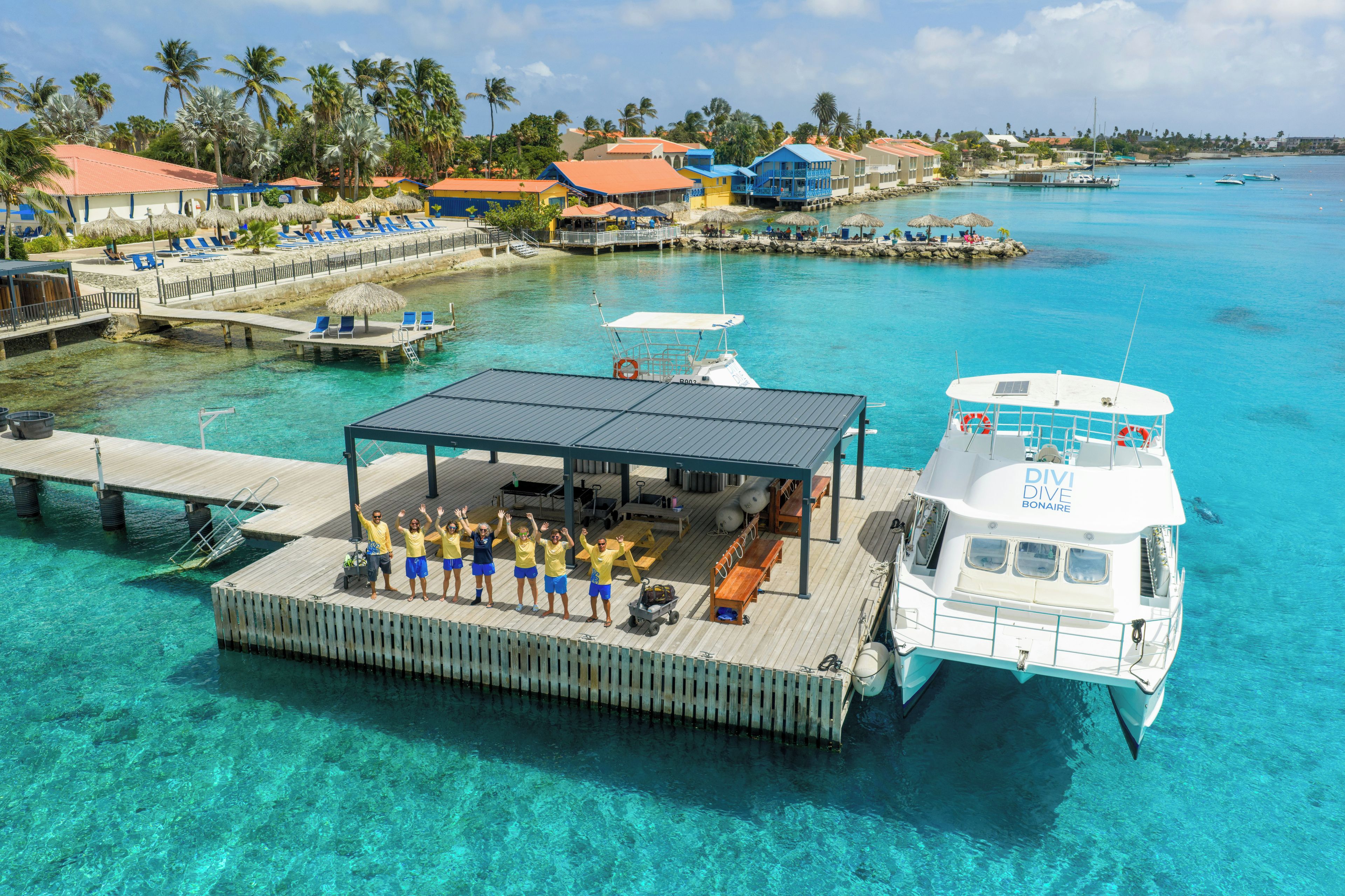 A boat docked at a pier with clear turquoise water, a covered deck with seating, and a backdrop of tropical buildings and palm trees.