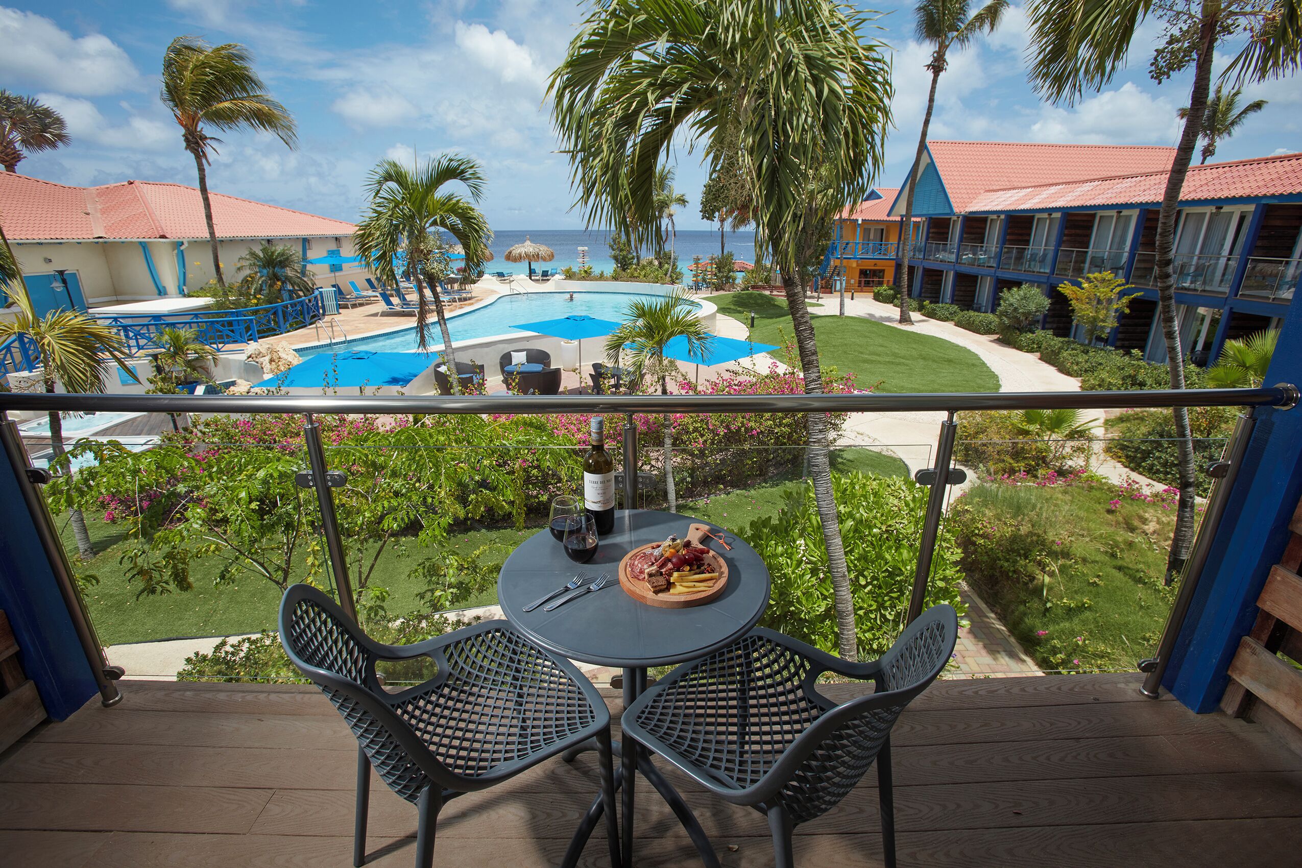 A balcony with a table set for two, overlooking a swimming pool and a resort building.