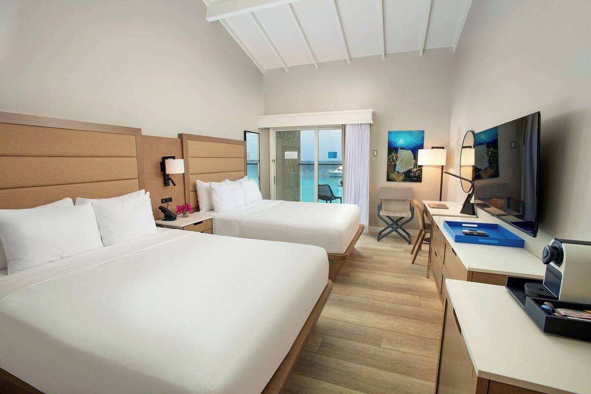 Habitación de hotel con dos camas, un escritorio, una silla, un televisor y una puerta corrediza a un balcón.