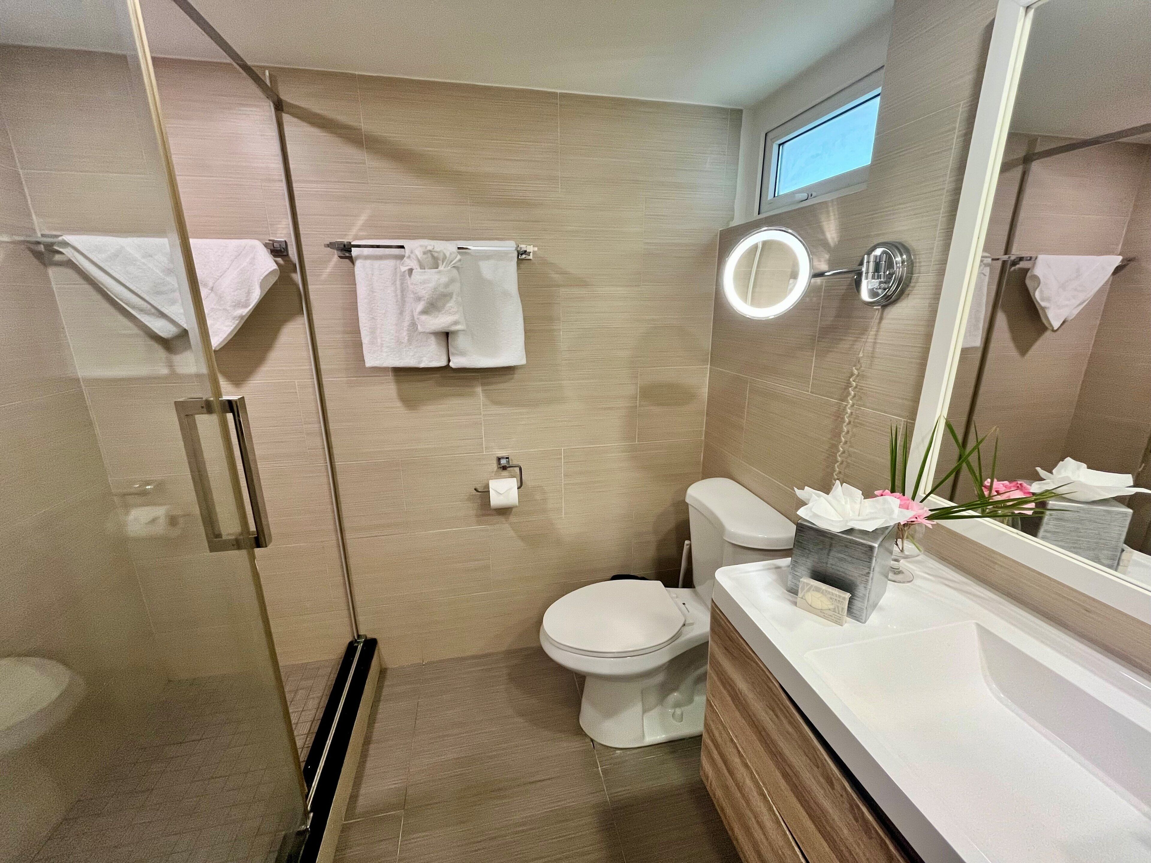 A modern bathroom with a glass shower enclosure, a toilet, a sink with a mirror, and a small window above the toilet.