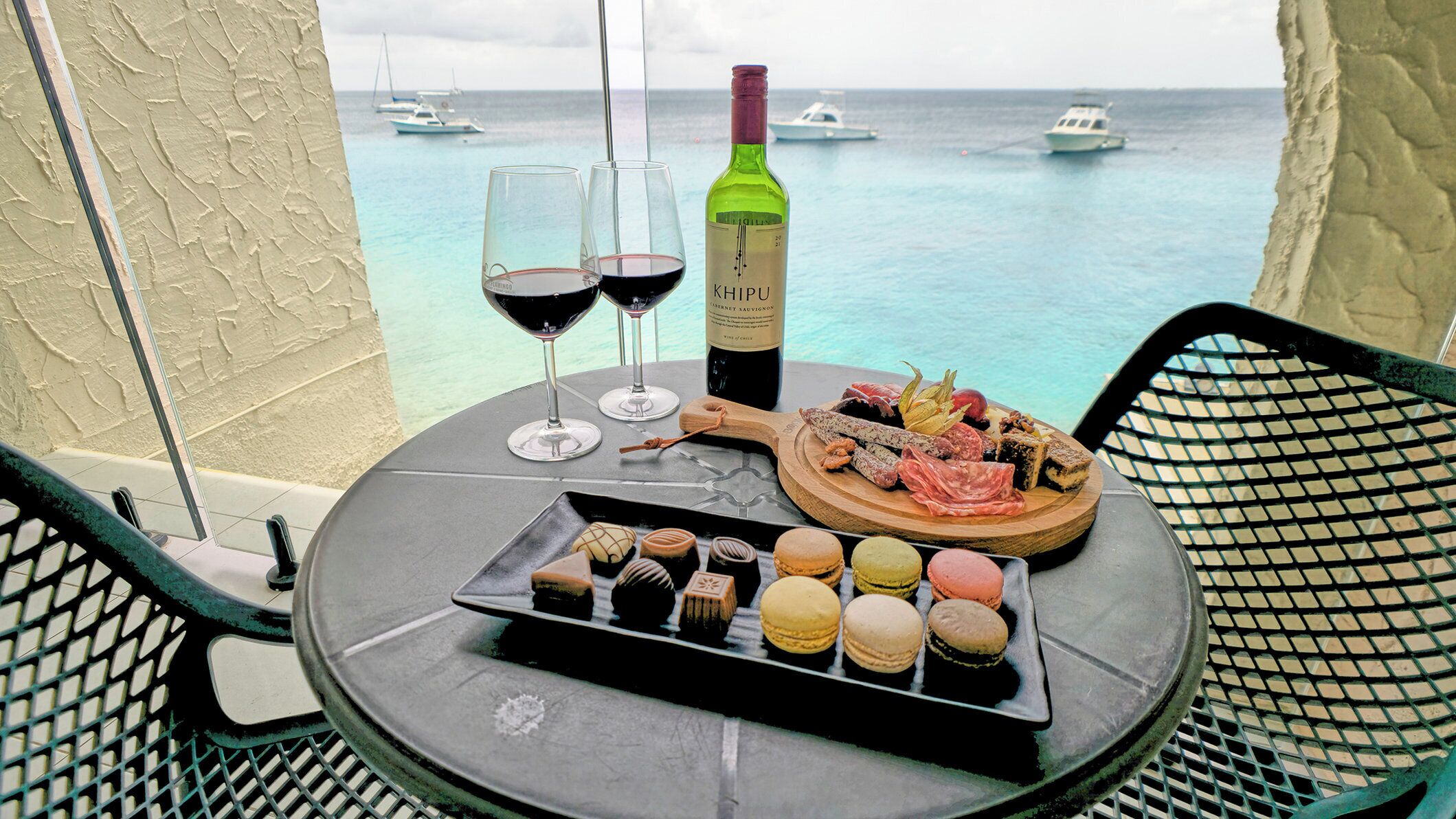 A table with a bottle of wine, glasses, a charcuterie board, and macarons on a balcony overlooking the sea.