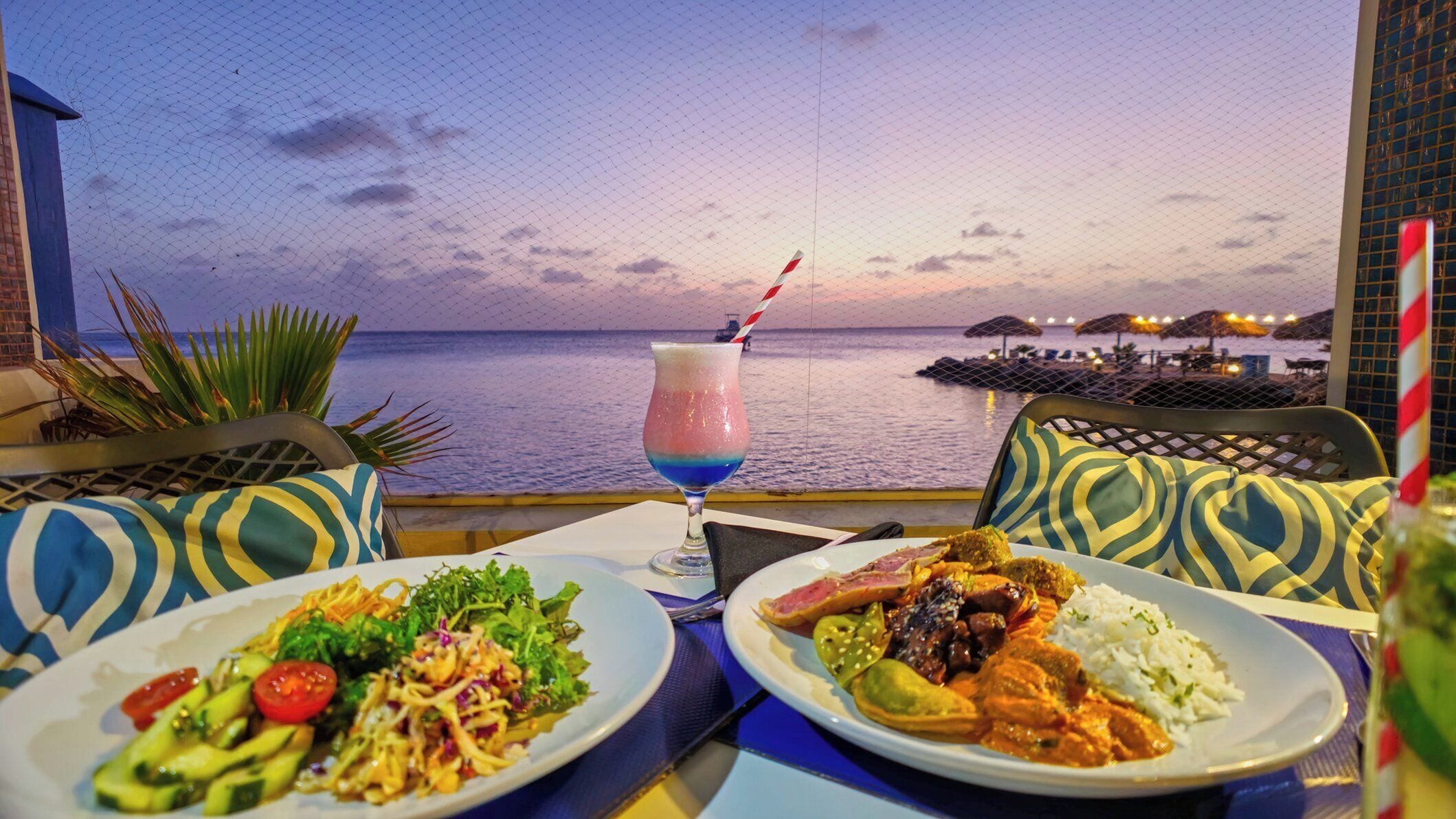 A table with two plates of food, a drink, and a view of the sea at sunset.