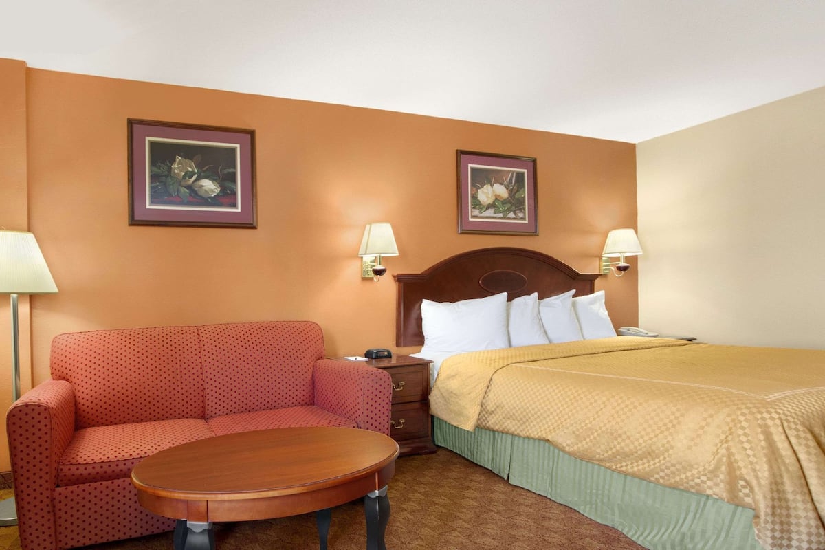 A hotel room with a sofa, a round table, a bed with a yellow bedspread, two framed floral paintings on the walls, and two bedside lamps.