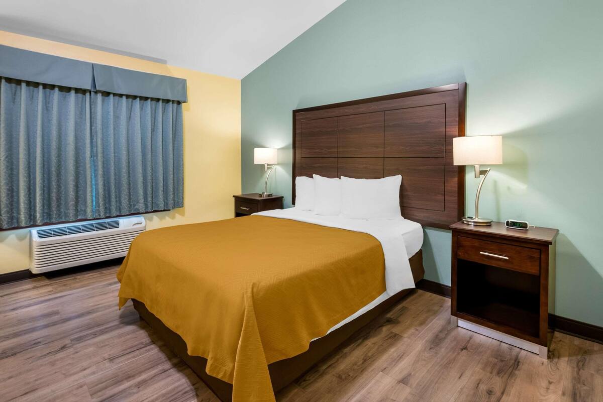 A hotel room with a bed, bedside tables, a lamp, an air conditioner, and a window with curtains.