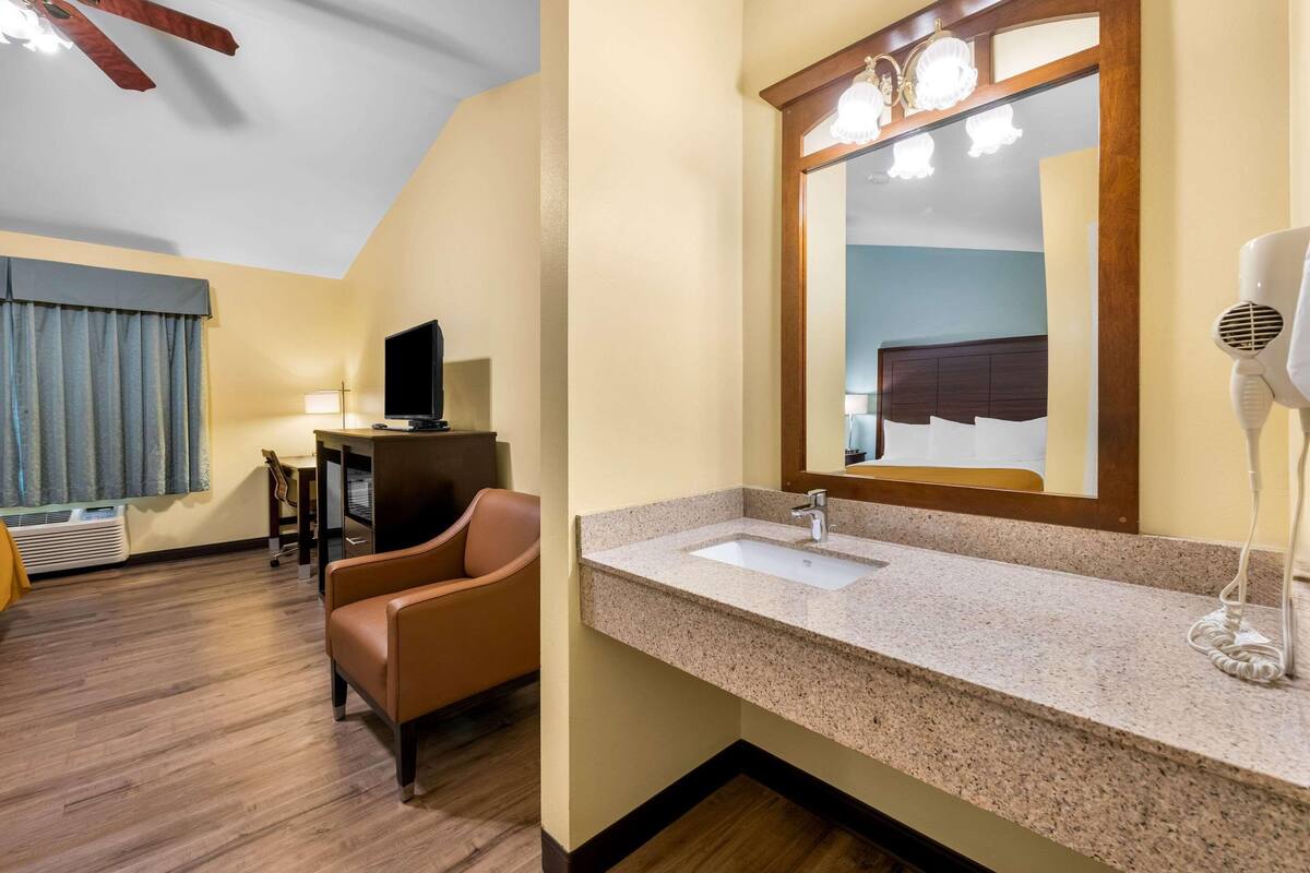 A hotel room with a large mirror, a sink, a hairdryer, a television, a chair, and a desk.