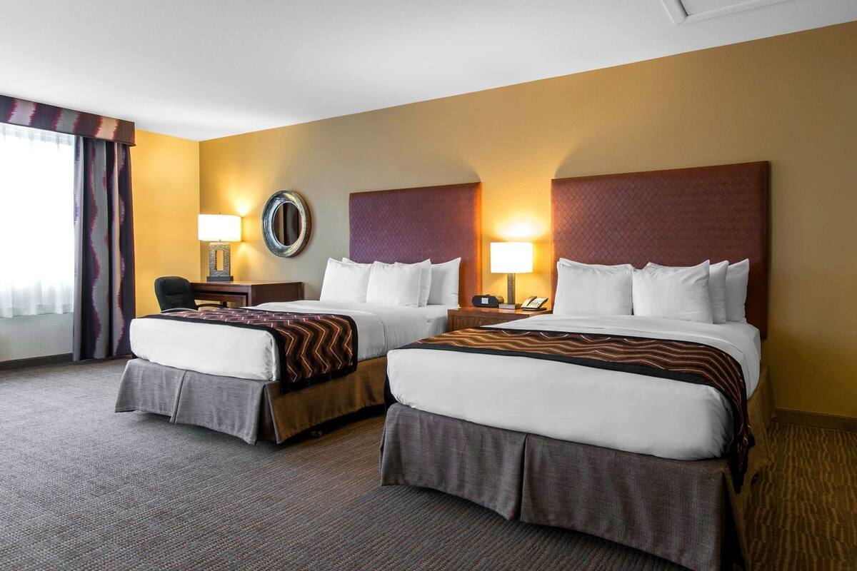 A hotel room with two beds, a desk, a chair, a lamp, and a large window with curtains.