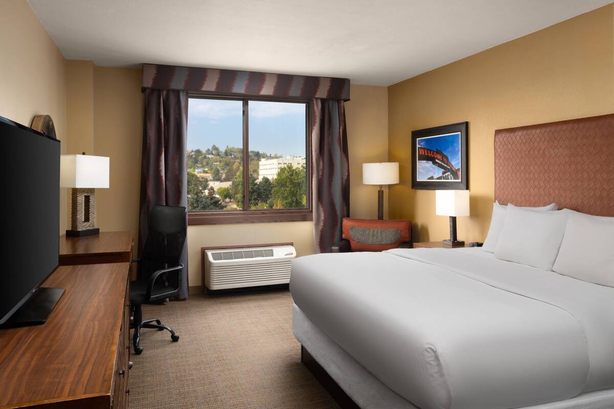 A hotel room with a large bed, a desk with a computer, a chair, a TV, a window with curtains, a wall-mounted heater, and a framed picture.