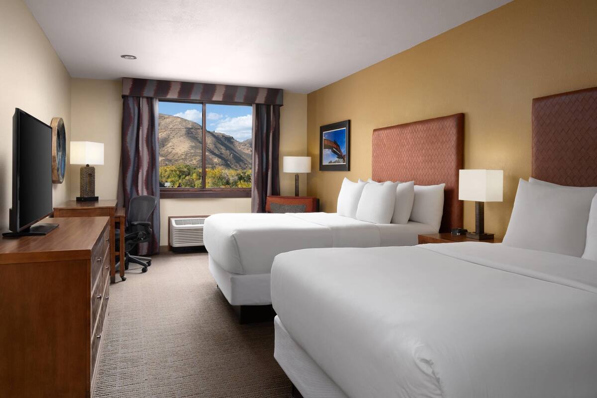 A hotel room with two beds, a desk, a television, and a window with a mountain view.