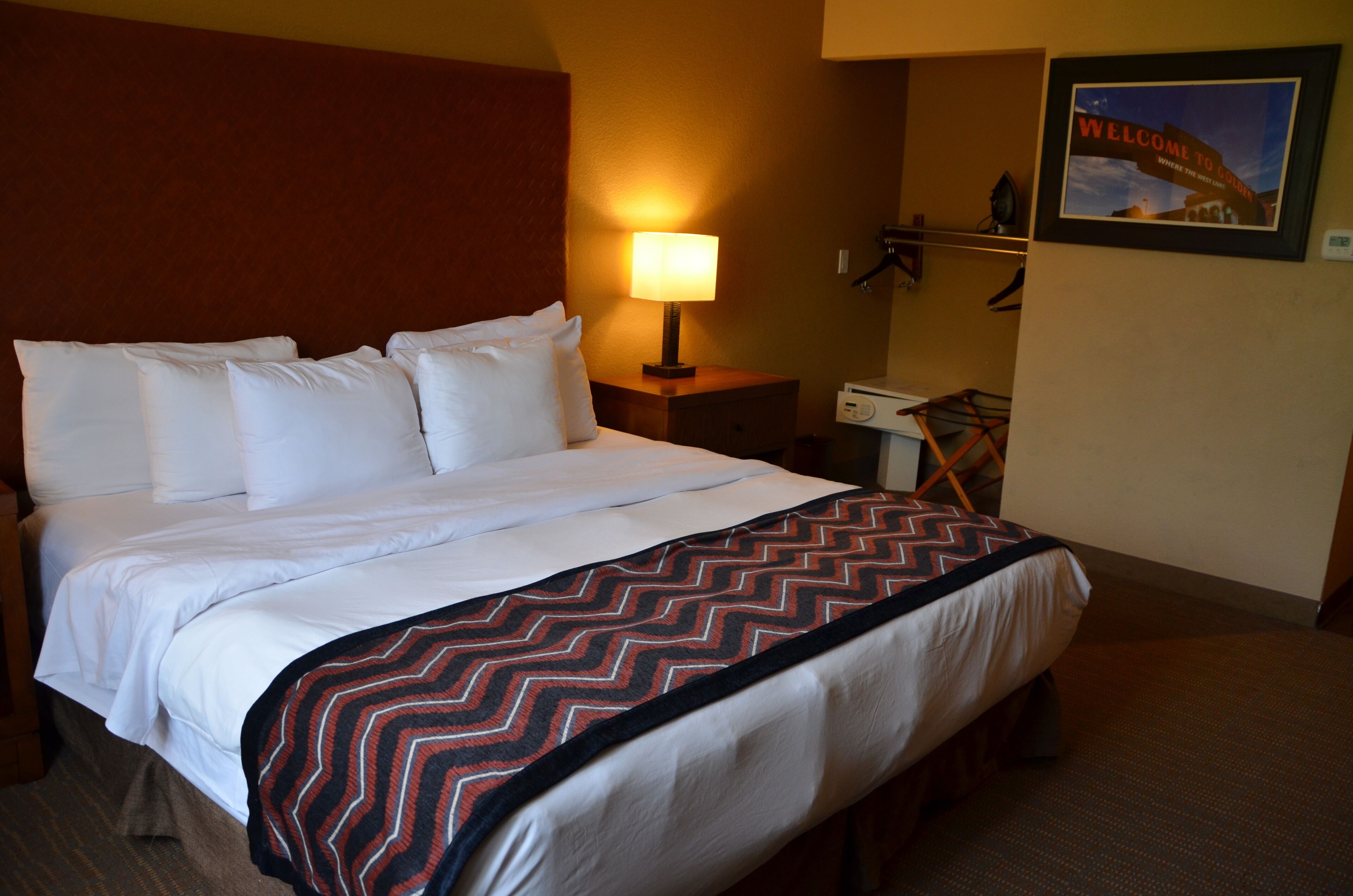 A hotel room with a large bed, a nightstand with a lamp, a desk with a telephone, and a welcome sign on the wall.