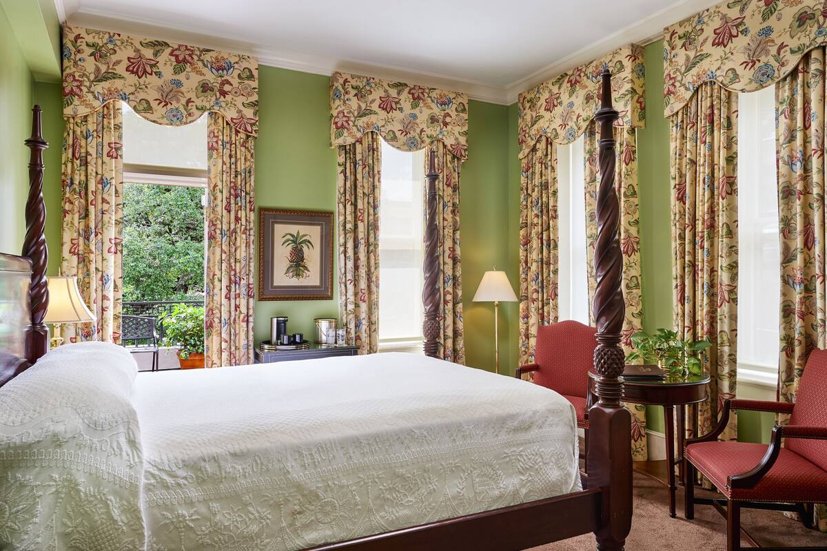 Une chambre avec un grand lit, des rideaux à motifs floraux, une chaise en bois, une petite table et une vue sur la verdure à l’extérieur.