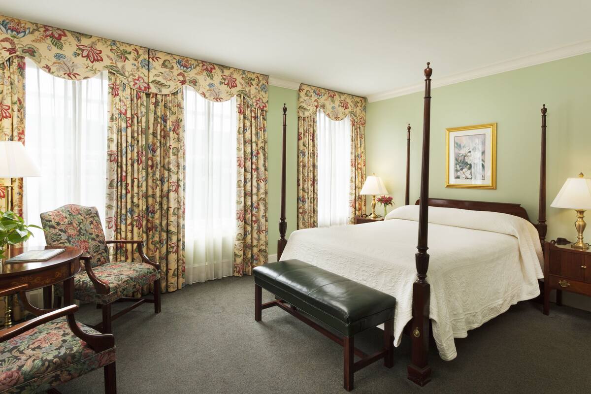 Un lit à baldaquin avec une couvre-lit blanche, un banc en bois, deux chaises recouvertes d’un tissu à motifs floraux et une table avec une lampe.