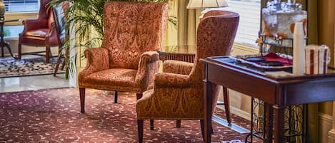 A room with a patterned carpet, a large armchair, a smaller armchair, a table with a lamp, and a potted plant.