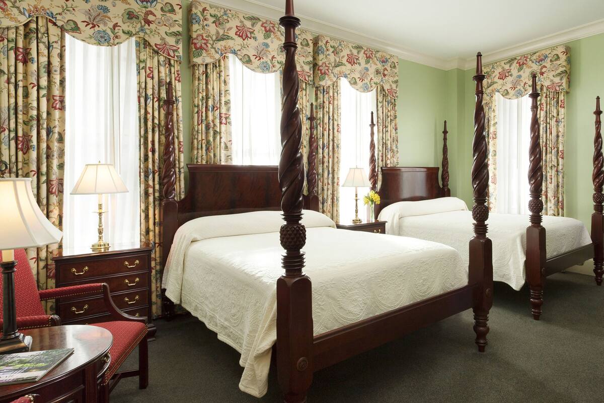 Un lit à baldaquin orné d’un motif floral, une table de chevet en bois avec une lampe, un fauteuil rembourré de couleur rouge et une commode à tiroirs.