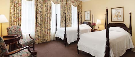 A hotel room with two single beds, floral curtains, a wooden chair, and a bedside table with a lamp.