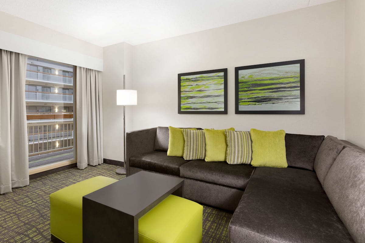 Habitació d'hotel moderna amb un sofà modular gris, una taula de cafè petita negra i un otoman verd brillant. Hi ha dos quadres abstractes emmarcats a la paret i un finestral gran amb cortines.