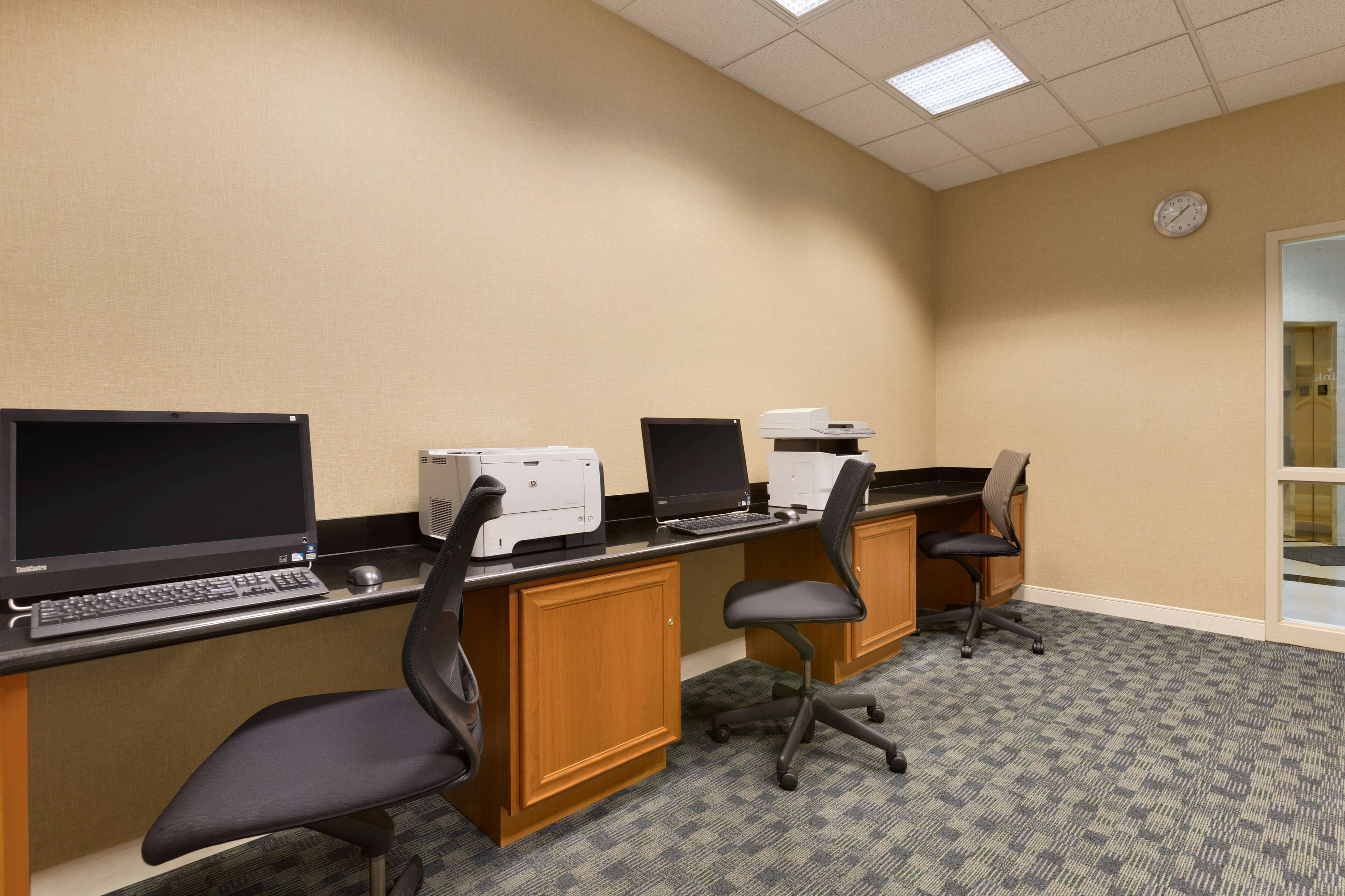 A hotel room with a desk, two computer monitors, a printer, and a clock on the wall.