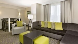 A modern hotel room with a grey sofa, a small table, and a dining area with chairs.