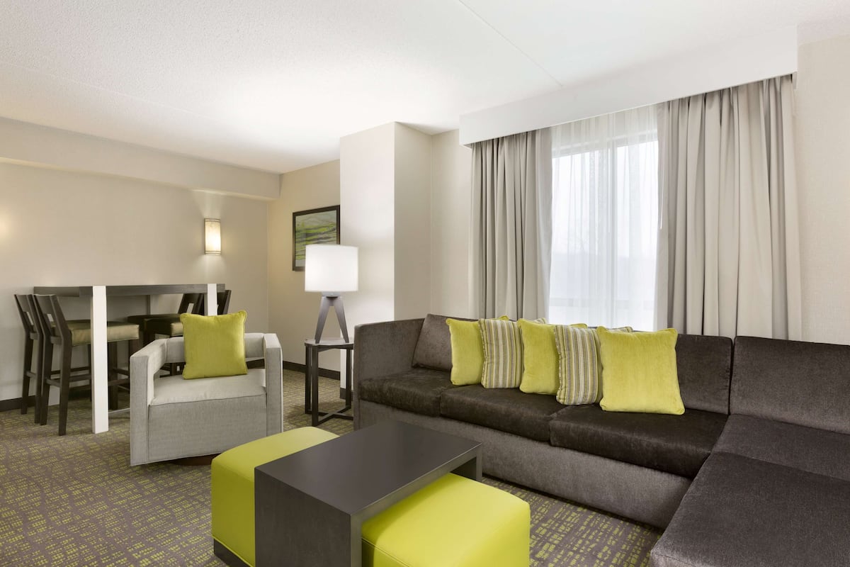 Habitació d'hotel moderna amb un sofà gris, una taula petita i zona de menjador amb cadires.